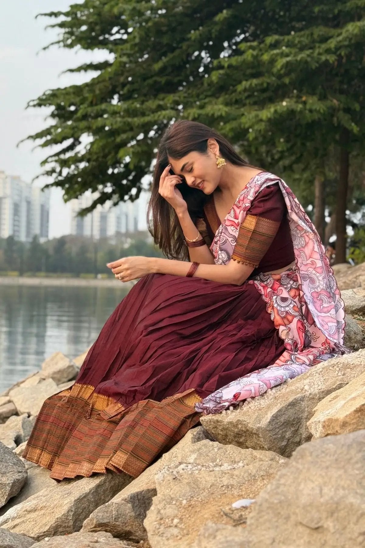 Model wearing Saumya Handloom Lehenga Set in maroon, seated outdoors by a lake, highlighting the flowing skirt and vibrant dupatta.