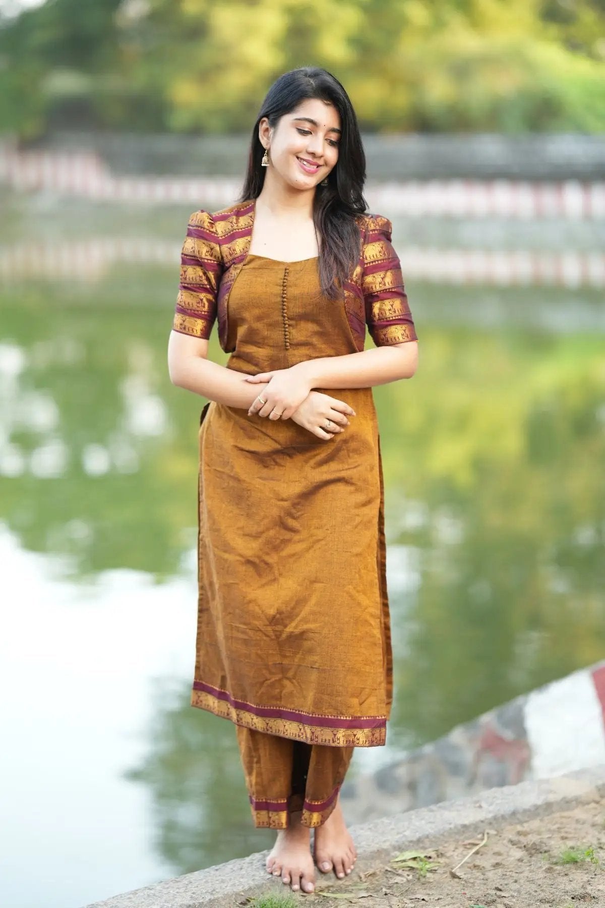 Barkha Kurta Set in ochre yellow with intricate maroon and gold patterned sleeves, styled for a traditional and elegant look near a serene lake.