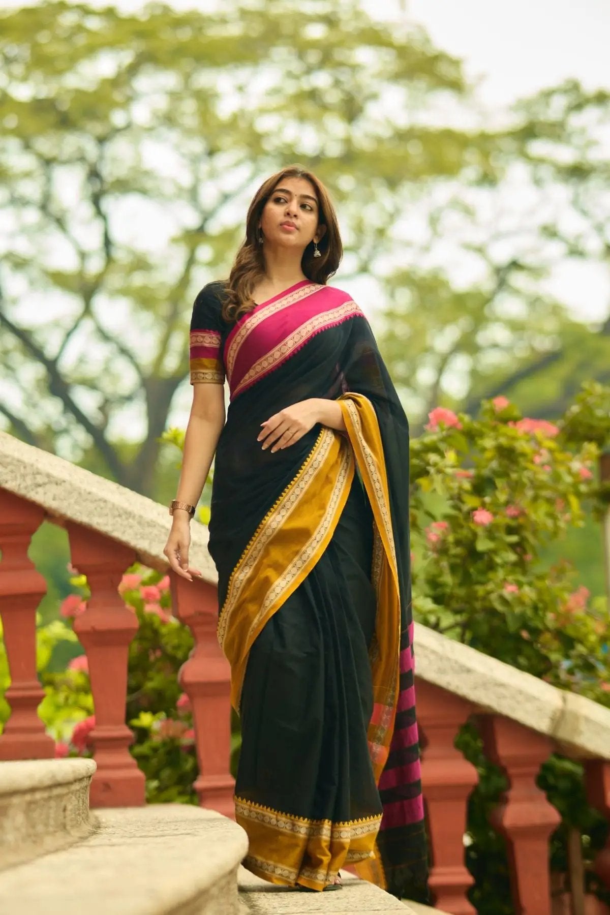 Black Gomti Handloom Saree with pink and gold border, draped gracefully by a model amidst greenery and architectural elements.