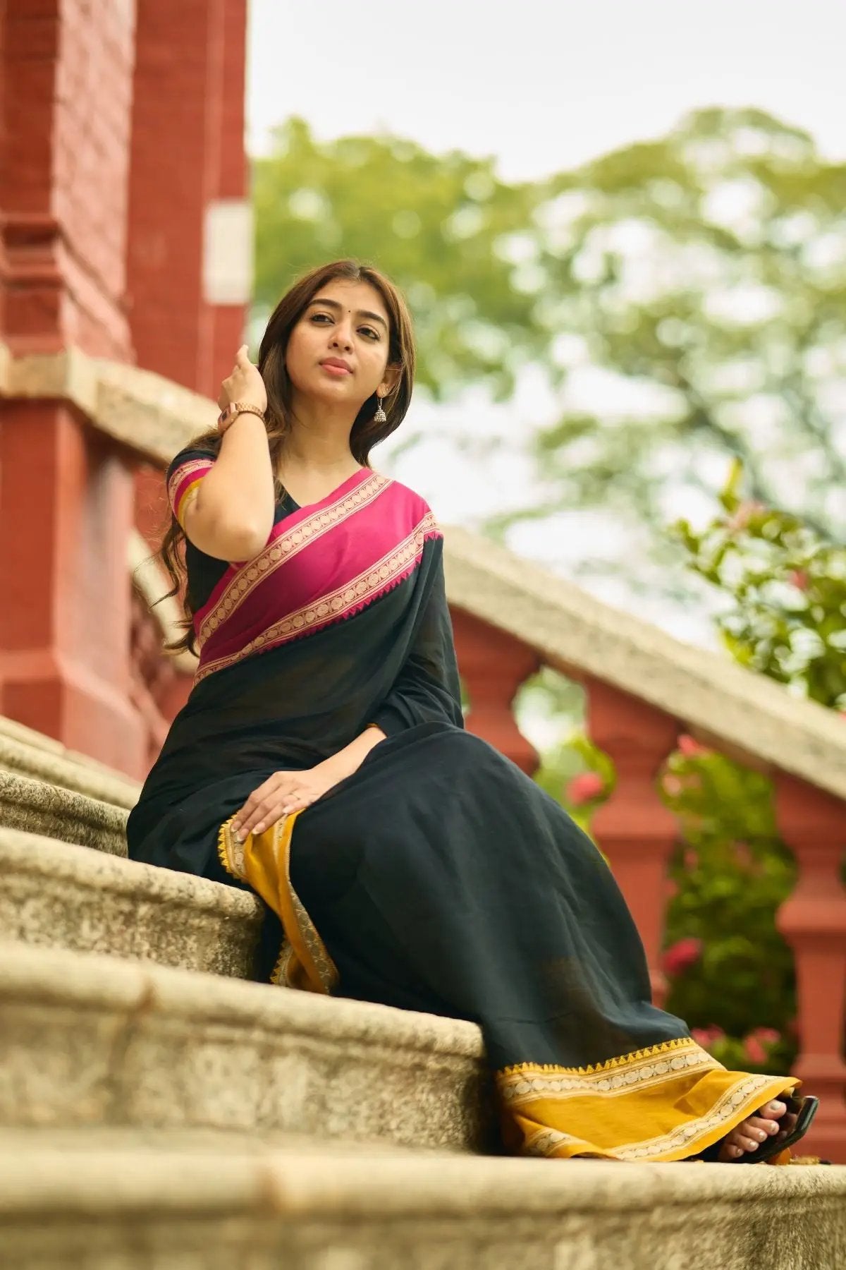 Traditional Gomti Handloom Saree in black with intricate pink and gold detailing, showcased by a model seated on stairs.