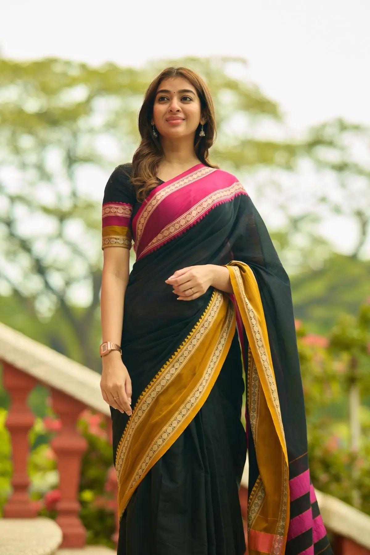 Exclusive Gomti Handloom Saree in black with vibrant pink and gold accents, worn by a model in a garden setting.