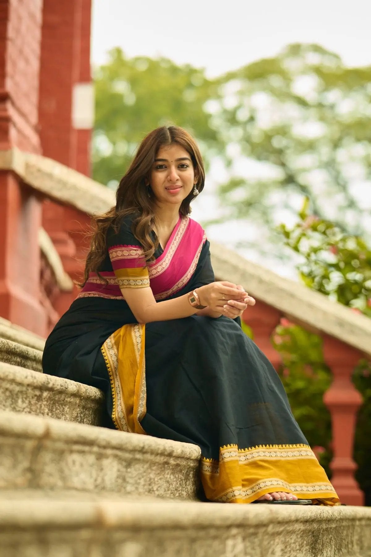 Elegant Gomti Handloom Saree in black with pink and gold accents, modeled outdoors on a staircase with a serene backdrop.