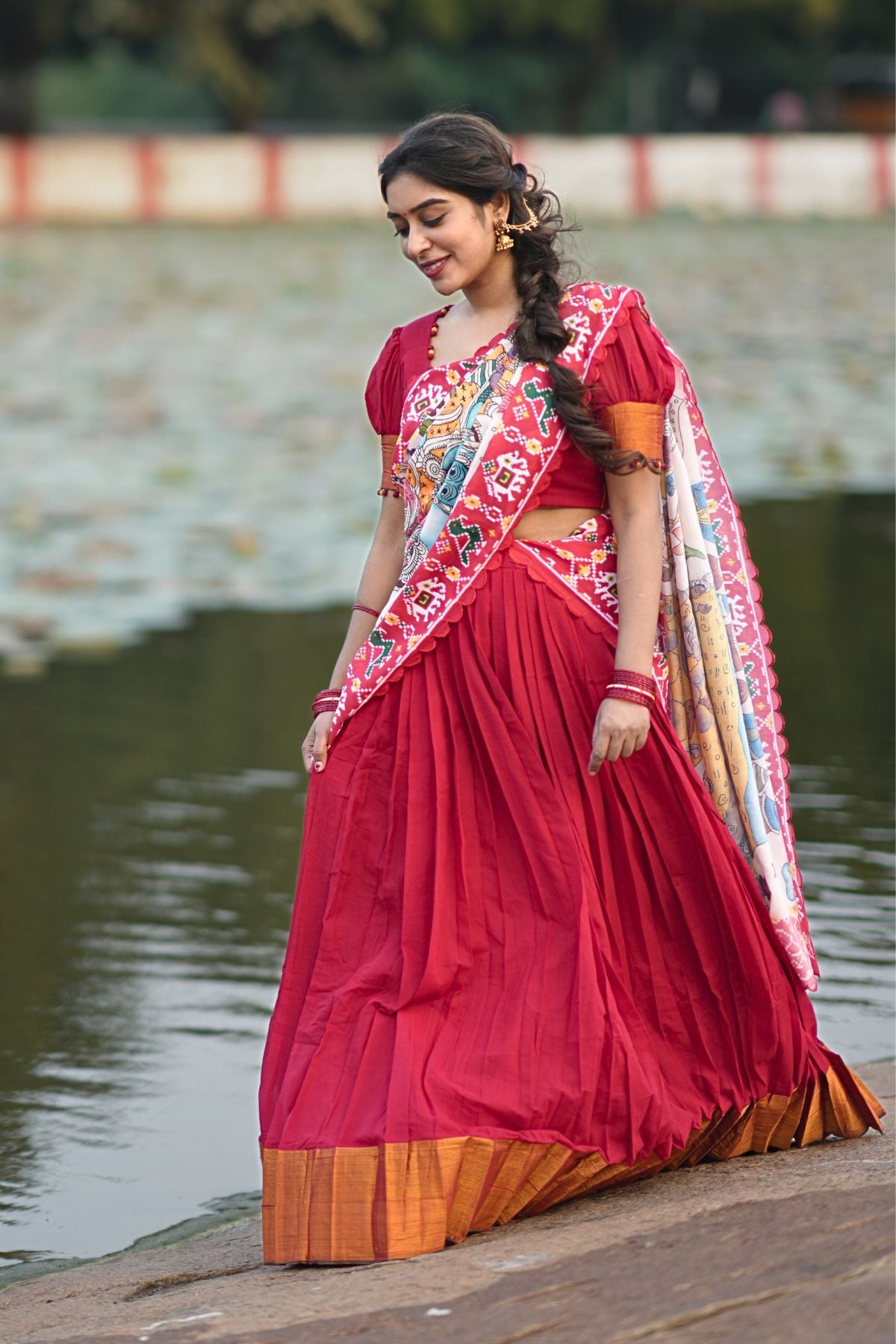 Revti Handloom Lehenga Set in Red with intricate embroidery and a vibrant multicolor dupatta, styled near a serene water body.