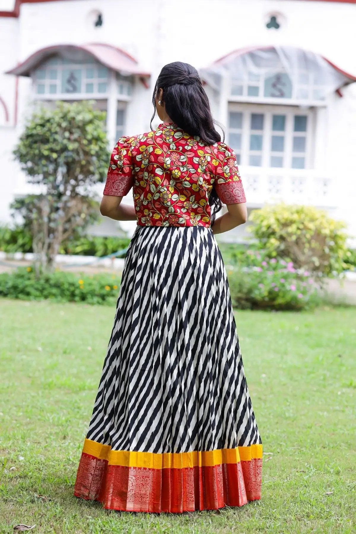 Back view of the Rohini Kalamkari Jacket Dress featuring a red floral jacket and striped black-and-white skirt with colorful borders in a garden setting.