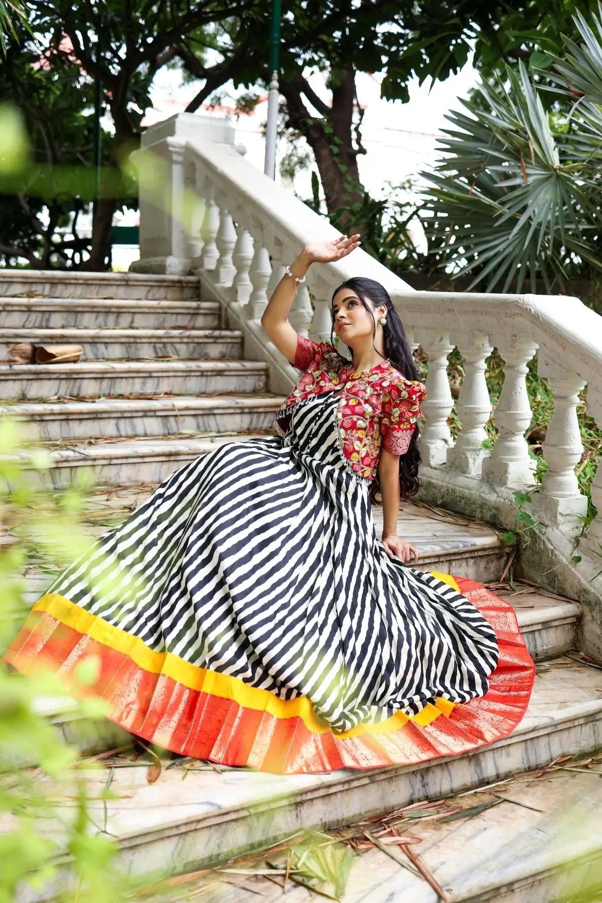 Stylish Rohini Kalamkari Jacket Dress in black with a vibrant red floral jacket and striped skirt, modeled on a marble staircase with greenery.